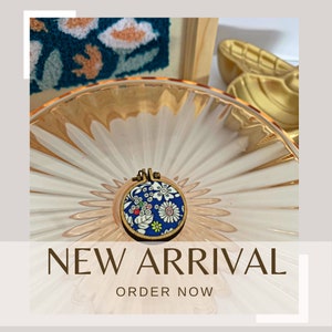 May include: A small, round embroidery hoop with a blue background and a floral pattern in white, red, green, and yellow. The hoop is on a white and gold background. "NEW ARRIVAL" and "ORDER NOW" are written in brown text below the hoop.