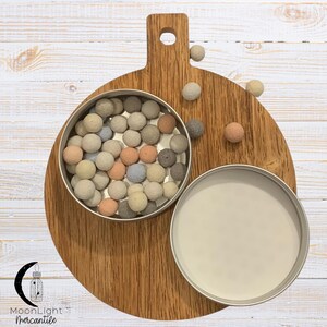 May include: A round metal tin filled with small, colorful, spherical objects in shades of beige, pink, blue, and gray. The tin sits on a wooden cutting board with a handle. Several spheres are scattered around the tin.