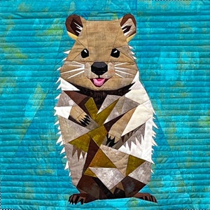 Könnte beinhalten: Ein gequilteter Wandbehang mit einem bunten Patchwork-Design eines lächelnden Quokkas, einem kleinen Beuteltier, das auf Rottnest Island, Australien, beheimatet ist. Das Quokka ist in einem hellbraunen, beigefarbenen und weißen Farbschema vor einem türkisblauen Hintergrund dargestellt.