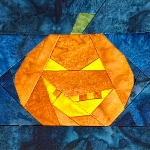 May include: A patchwork pumpkin with a carved jack-o'-lantern face. The pumpkin is made of orange and yellow fabric pieces, and the face is outlined in black. The background is a dark blue fabric.
