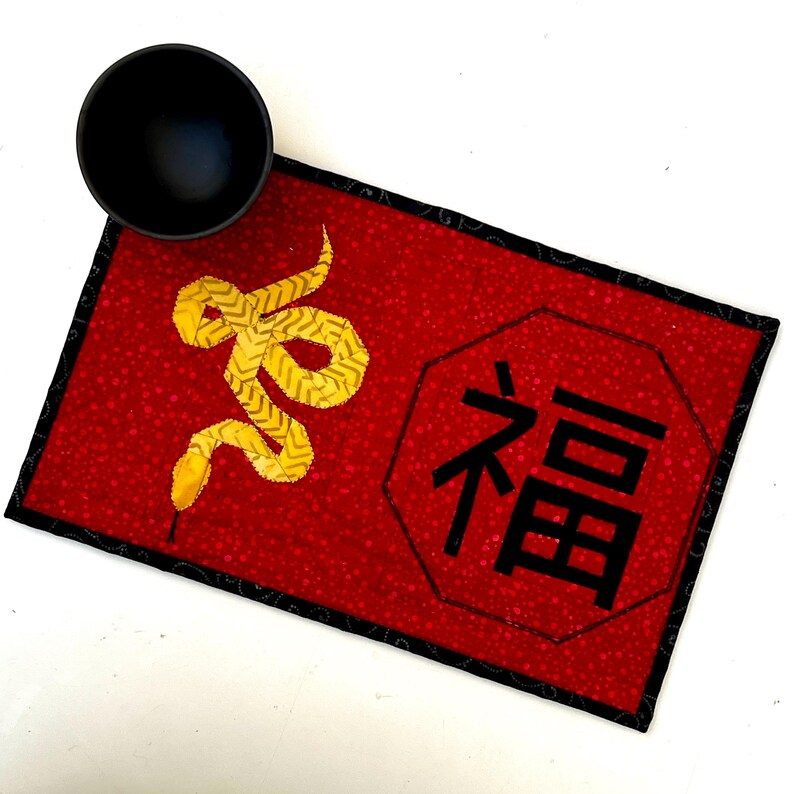 May include: Red fabric placemat with a black border and a gold snake design. The placemat also features a black octagon with the Chinese character for "fortune" in white.