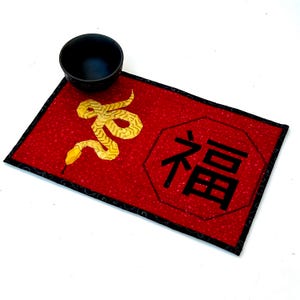 May include: Red fabric placemat with a black border and a gold snake design. The placemat also features a black octagon with the Chinese character for "good fortune" in white.