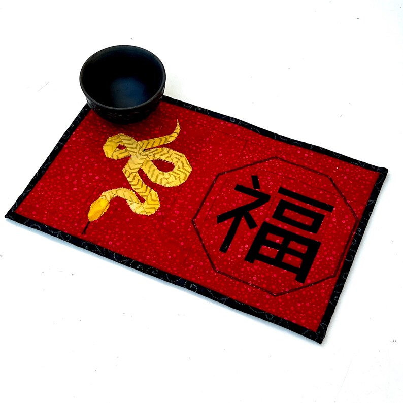 May include: Red fabric placemat with a black border and a gold snake design. The placemat also features a black octagon with the Chinese character for "good fortune" in white.