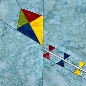May include: A colorful patchwork quilt block featuring a kite design with yellow, red, blue, and green fabric pieces. The kite is set against a light blue background.