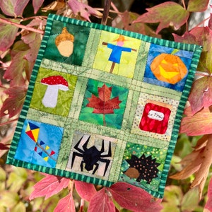 Könnte beinhalten: Ein farbenfroher Patchwork-Quiltblock mit neun Quadraten, die herbstliche Themen zeigen. Die Quadrate zeigen einen Vogelscheuche, einen Kürbis, einen Pilz, ein Ahornblatt, ein Glas Marmelade, einen Drachen, eine Spinne, einen Igel und eine Eichel.