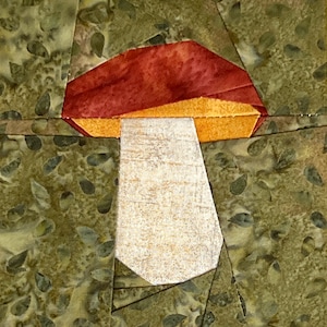 Peut inclure: Une applique de champignon rouge et blanc avec un pied brun, réalisée à partir de chutes de tissu. Le champignon est placé sur un fond vert avec un motif de feuilles.