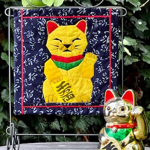 May include: A yellow and black Maneki Neko cat quilt with a red border is displayed on a metal stand. A gold-coloured Maneki Neko figurine sits to the right. The quilt features a yellow cat holding a gold coin with Japanese characters.