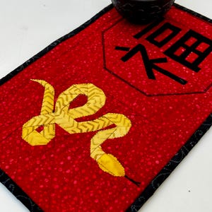May include: A red fabric coaster with a black border and a gold snake design. The coaster also features a black octagon with the Chinese character for "snake" in black.