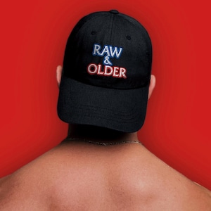 May include: Black baseball cap with the embroidered text "RAW & OLDER" in blue and red. The cap is worn backward against a red background.