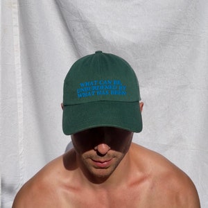 May include: A dark green baseball cap featuring the blue embroidered text: "WHAT CAN BE, UNBURDENED BY WHAT HAS BEEN". The cap has a curved peak and is made of a soft material. A fashionable headwear item.
