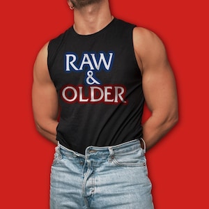 以下が含まれることがあります： 白、青、赤で「RAW & OLDER」の文字が入った黒いノースリーブシャツ。明るいブルーデニムのジーンズと合わせています。背景は無地の赤色です。