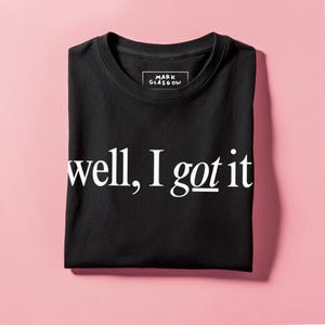 Könnte beinhalten: Schwarzes T-Shirt mit dem weißen Text "well, I got it". Der Markenname "MARK GLASGOW" steht auf dem Etikett. Das T-Shirt liegt gefaltet auf einem rosa Hintergrund.