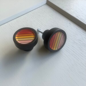 May include: Two black round cabinet knobs with a colourful striped design. The stripes are orange, yellow and pink.