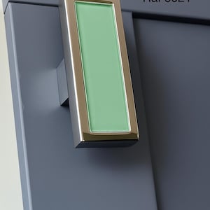 May include: A rectangular cabinet pull with a polished chrome finish and a light green glass insert. The pull is mounted on a gray cabinet door. The text "Ral 6021" is visible on the door.