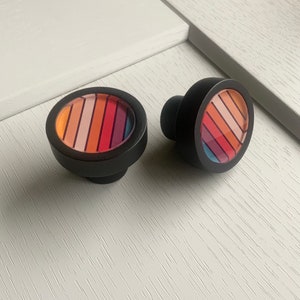 May include: Two black round cabinet knobs with a colorful striped design. The stripes are in shades of orange, pink, red, and purple.