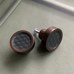 Könnte beinhalten: Zwei bronzefarbene Schrankknöpfe. Jeder Knopf hat eine kreisförmige Oberfläche mit dunklem Hintergrund und einem sich wiederholenden Wellenmuster in einer helleren Farbe. Die Knöpfe sind auf einer hellgrünen Oberfläche montiert.