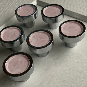 May include: Six chrome cabinet knobs with a pink marble design. The knobs are round and have a raised edge.