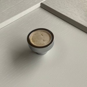 May include: A round, chrome knob with a beige centre. The knob is designed for use on cabinets or drawers.