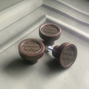 May include: Three bronze cabinet knobs with a gold geometric pattern. The knobs have a rounded shape with a decorative face and a short stem. The hardware is designed for furniture and home decor.