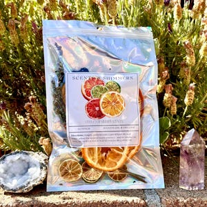 May include: A clear, iridescent bag of "SCENT SHIMMERS" stovetop simmer scents, featuring dried citrus slices. The label reads "SUMMER, HANDMADE & ORGANIC." A geode and a purple crystal are next to the bag. Lavender and other plants are in the background.