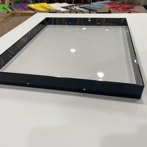May include: A black frame with a clear glass insert. The frame is sitting on a white surface.