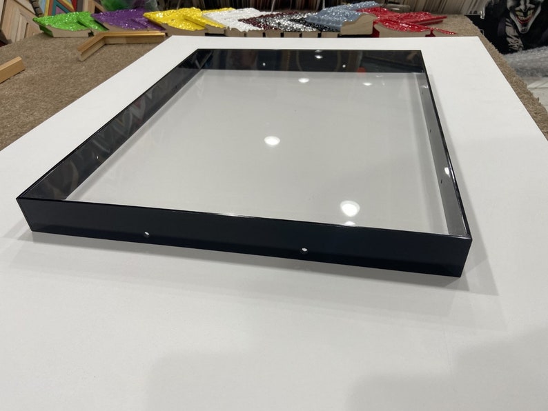 May include: A black frame with a clear glass insert. The frame is sitting on a white surface.