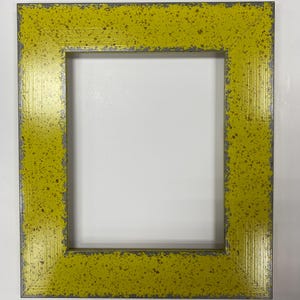 May include: A rectangular picture frame with a distressed yellow finish and gray accents. The frame has a speckled pattern and a textured surface, creating a vintage or rustic aesthetic. The frame is designed to hold a photograph or artwork.