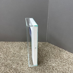 Op de afbeelding: Een transparante acryl display case met een wit interieur en zichtbaar kunstwerk. De behuizing is rechthoekig en heeft een glasachtige uitstraling. Het kunstwerk binnenin is gedeeltelijk zichtbaar, met blauwe en witte tinten.