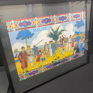 May include: A framed illustration of a beach scene with people in colorful clothing. The illustration is titled "MALIBU" and features a palm tree, a dog, and a beach umbrella. The illustration is framed in a black frame.