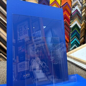 May include: A blue display stand with clear acrylic panels. The stand is designed to hold framed artwork or photographs. The background features a variety of colorful picture frames, suggesting a framing shop or art gallery.