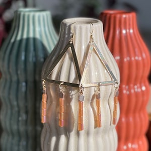 Puede incluir: Pendientes de triángulo dorado con cuentas colgantes de color melocotón. Los pendientes se muestran en un jarrón blanco texturizado.