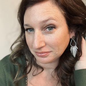 Puede incluir: Una mujer con el pelo largo y castaño que lleva un jersey verde y pendientes de aro plateados. Los pendientes tienen un pequeño círculo en la parte superior y una franja de finas hebras plateadas.