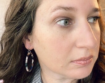 May include: A woman with long brown hair is wearing a pair of large, oval-shaped hoop earrings. The earrings are made of a patterned material with shades of blue, white, and black.
