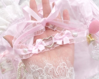 May include: Pink lace choker with a silver heart charm and two pink bows. The choker is on a hand with a white lace background.