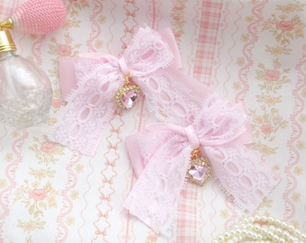 Può includere: Due fiocchi di raso rosa con bordino in pizzo e dettagli in cristallo. I fiocchi sono su un tessuto a fantasia floreale rosa e bianco.