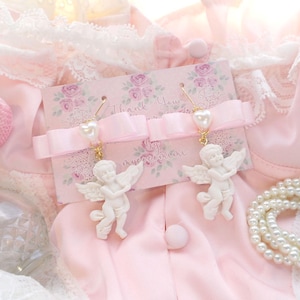 K&ouml;nnte beinhalten: Ein Paar wei&szlig;e Cherub-Ohrringe mit Perlenakzenten. Die Ohrringe werden auf einer rosa Karte mit dem Text "Thank You" und "by mysecretmini" pr&auml;sentiert.
