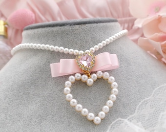 Peut inclure: Un collier délicat de perles avec un ruban rose et un pendentif en forme de cœur orné d'un strass rose. Le collier est posé sur un tissu de velours gris.