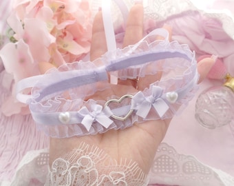 May include: A lavender lace garter with a silver heart charm and pearl accents. The garter is tied with a bow and is held in a hand.