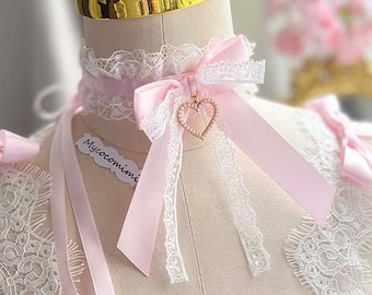 Puede incluir: Un top tipo corsé rosa claro con ribetes de encaje blanco, lazos de cinta de satén y un colgante en forma de corazón. La prenda incluye una gargantilla de encaje a juego con un lazo rosa y un pequeño dije de corazón. El texto Mycocomimi es visible.