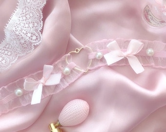 May include: A pink satin fabric background with a delicate lace trim. A pink choker with pearls and bows, a gold heart charm, and a vintage perfume bottle with a pink bulb atomizer are featured.
