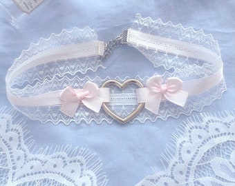 May include: A delicate white lace choker with a pink ribbon and a silver heart charm. The choker has two pink bows on either side of the heart charm.