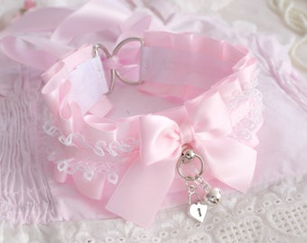 May include: A pink satin choker with a white lace trim and a bow. The choker has a silver charm with a heart and a bell.