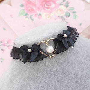 Może przedstawiać: Czarny choker z koronki z złotym sercem i perłowym wisiorem. Choker ma dwie czarne satynowe kokardki z perłowymi akcentami.