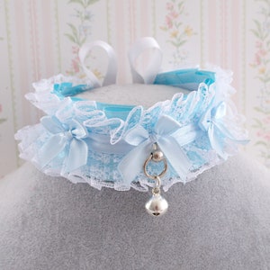 May include: A light blue lace choker with a silver bell charm and a silver ring with a bead. The choker has two white satin bows on each side.