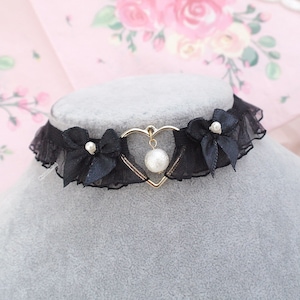 Może przedstawiać: Czarny choker z koronki z złotym wisiorem w kształcie serca i perłową zawieszką. Choker ma dwie czarne kokardki z perłowymi akcentami.