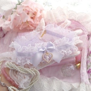 May include: A delicate white lace garter with a lavender bow and a pearl heart charm. The garter is on a pink background with other lace items.