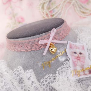 May include: A pink lace choker necklace with a small bow and a gold heart charm. A sticker of a white cat with a pink bow is next to the necklace. The word "Macaromi" is written in gold script on the sticker and on a separate piece.
