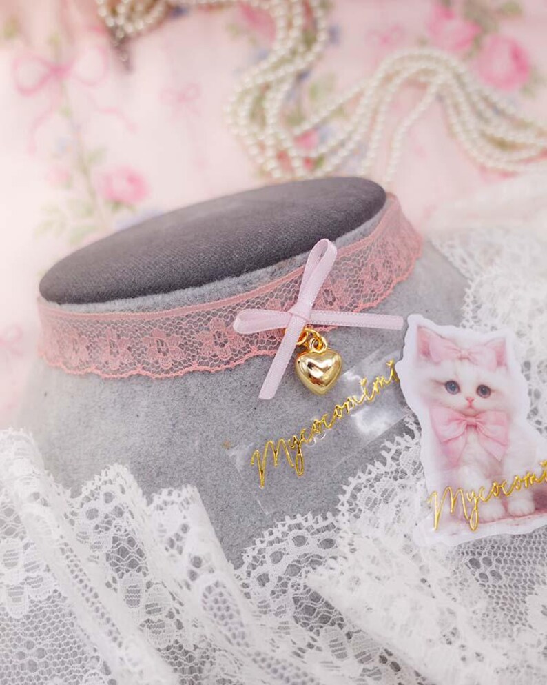 May include: A pink lace choker necklace with a small bow and a gold heart charm. A sticker of a white cat with a pink bow is next to the necklace. The word "Macaromi" is written in gold script on the sticker and on a separate piece.