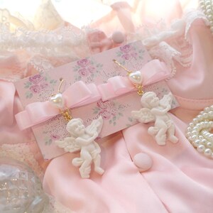 K&ouml;nnte beinhalten: Ein Paar wei&szlig;e Cherub-Ohrringe mit Perlenakzenten und goldfarbenen Haken. Die Ohrringe werden auf einer rosa Karte mit floralem Design und dem Text "Thank You" und "by muccacimeni" pr&auml;sentiert.