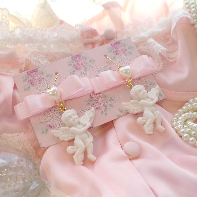 K&ouml;nnte beinhalten: Ein Paar wei&szlig;e Cherub-Ohrringe mit Perlenakzenten und goldfarbenen Haken. Die Ohrringe werden auf einer rosa Karte mit floralem Design und dem Text "Thank You" und "by muccacimeni" pr&auml;sentiert.